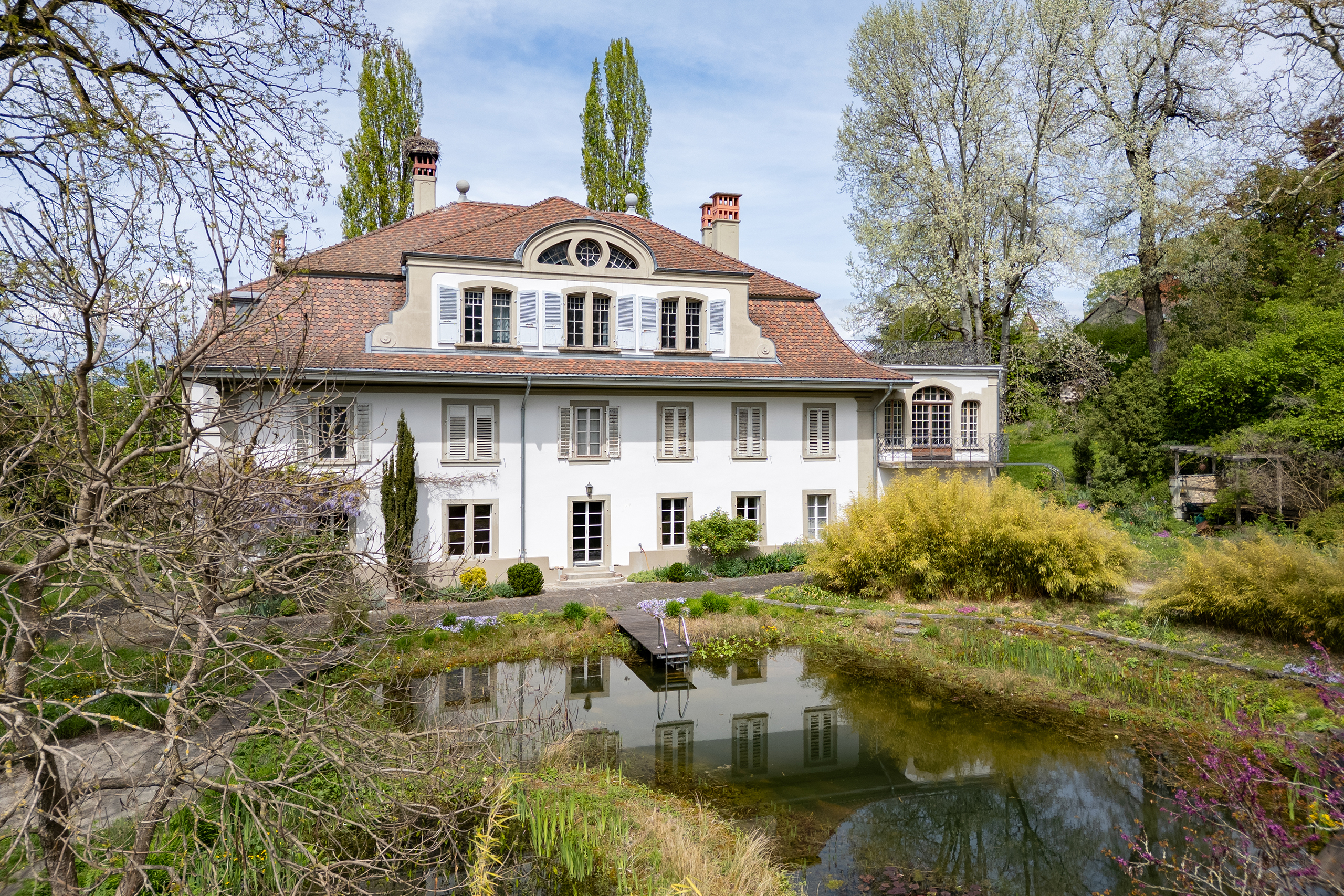 Drohnenaufnahme mit Sicht auf die Südseite eines Herrschaftshauses. Im Bildvordergrund steht der grosse Schwimmteich mit Steg.